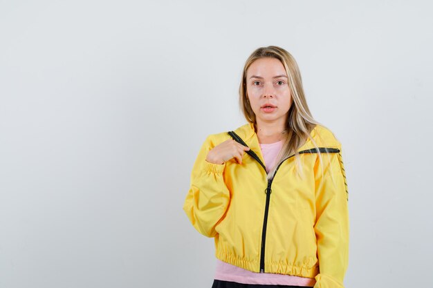 Blonde girl pointing at herself in yellow jacket and looking puzzled