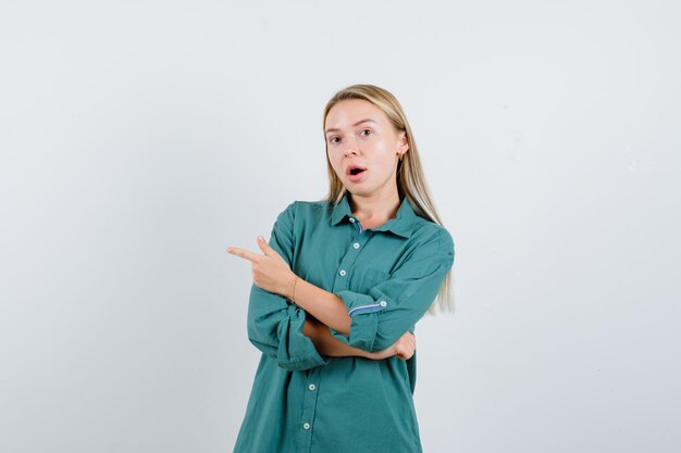 Блондинка, указывая влево указательным пальцем в зеленой блузке и выглядя удивленно.