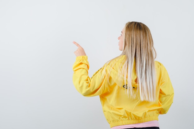 Blonde girl pointing left with index finger, turning back in pink t-shirt and yellow jacket and looking alluring