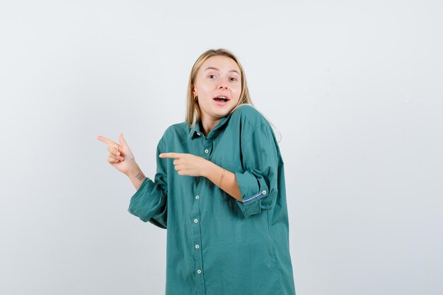 Blonde girl pointing left with index fingers in green blouse and looking happy