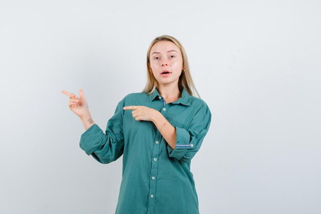 Blonde girl pointing left with index fingers in green blouse and looking serious,