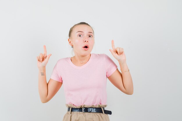 Blonde girl pointing up in t-shirt, pants and looking scared , front view.