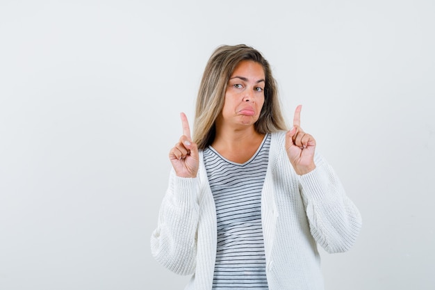 Ragazza bionda rivolta verso l'alto con il dito indice in maglietta a righe, cardigan bianco e pantaloni jeans e guardando sorpreso. vista frontale.