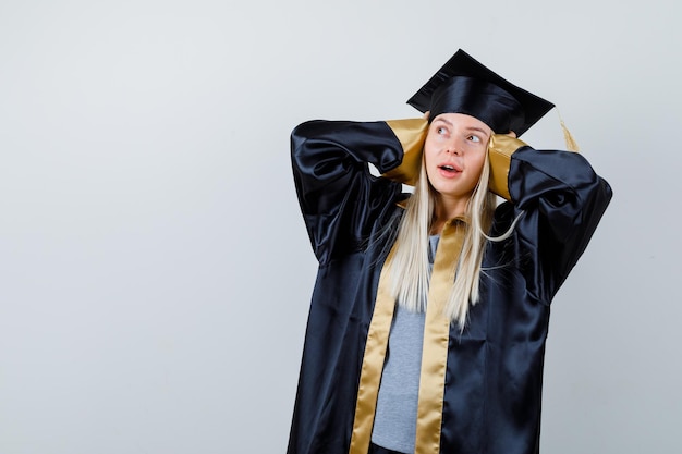 Blonde girl pressing hands on ears, looking at left side in graduation gown and cap and looking focused.