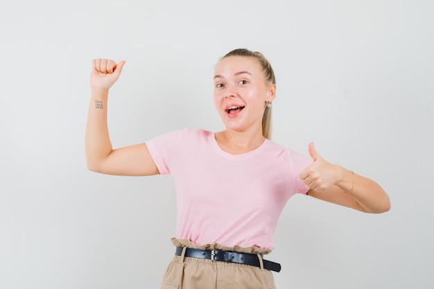 Blonde girl pretending to hold something, showing thumb up in t-shirt, pants , front view.