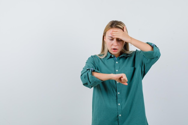 Free photo blonde girl pretending like looking at clock while putting hand on forehead in green blouse and looking surprised