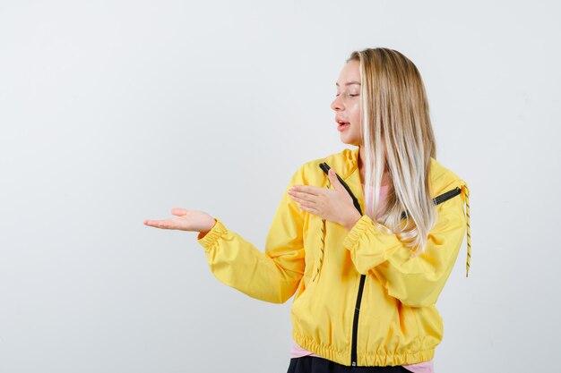 Blonde girl pretending to show something in yellow jacket and looking cute