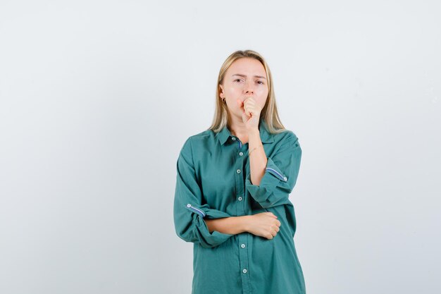 Blonde girl putting hand on mouth while holding hand on belly in green blouse and looking sleepy