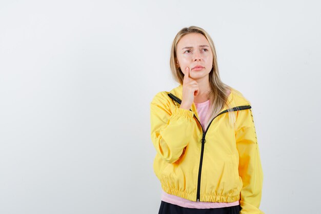 Blonde girl putting index finger near mouth, thinking about something in pink t-shirt and yellow jacket and looking pensive.