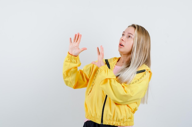 Blonde girl raising hands to defend herself in yellow jacket and looking scared