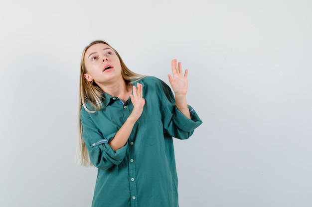 Blonde girl raising hands in surrender pose in green blouse and looking scared.