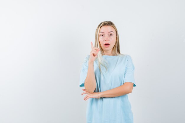 Blonde girl raising index finger in eureka gesture while holding hand under elbow in blue t-shirt and looking sensible , front view.