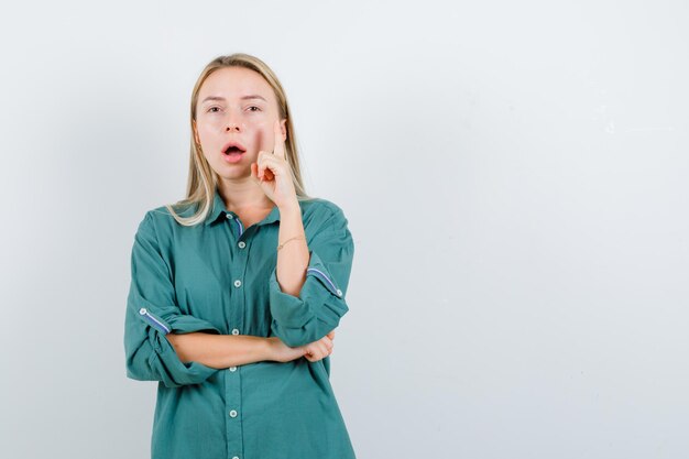 Blonde girl raising index finger in eureka gesture while holding hand on elbow in green blouse and looking sensible.