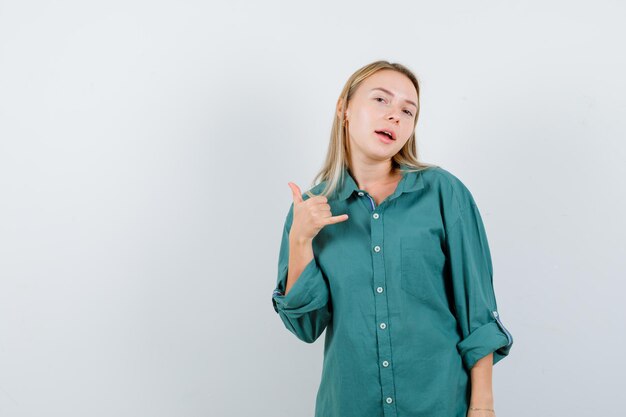 Blonde girl showing call me gesture in green blouse and looking happy.
