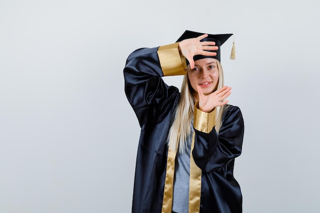 Free photo blonde girl showing camera gesture in graduation gown and cap and looking happy