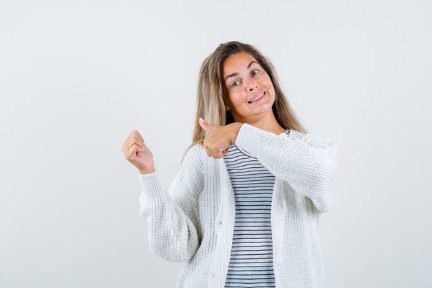 ストライプのTシャツ、白いカーディガン、ジーンズのズボンで二重の親指を見せて、幸せそうに見えるブロンドの女の子。正面図。