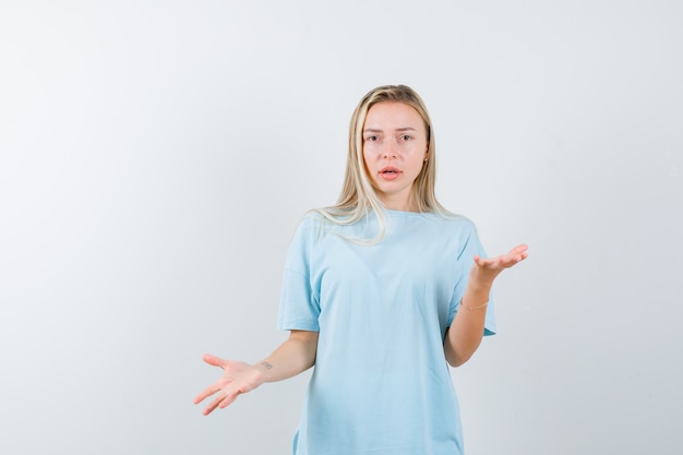 Ragazza bionda che mostra gesto impotente in maglietta blu e guardando perplesso, vista frontale.