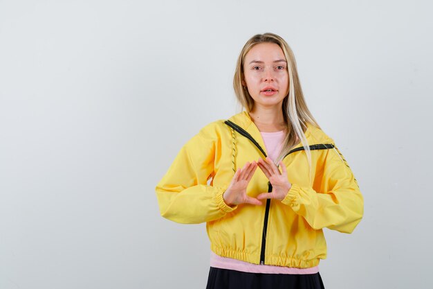 Blonde girl showing insurance gesture in pink t-shirt and yellow jacket and looking serious.