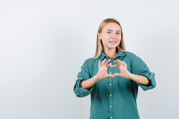 Blonde girl showing love gesture with hands in green blouse and looking enchanting