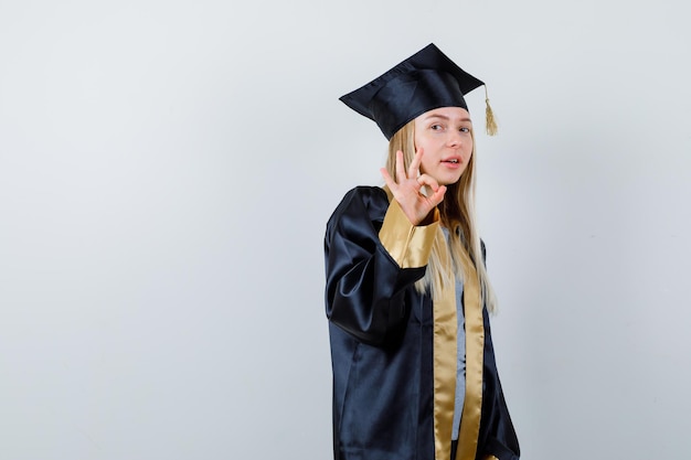 Blonde girl showing ok sign in graduation gown and cap and looking happy