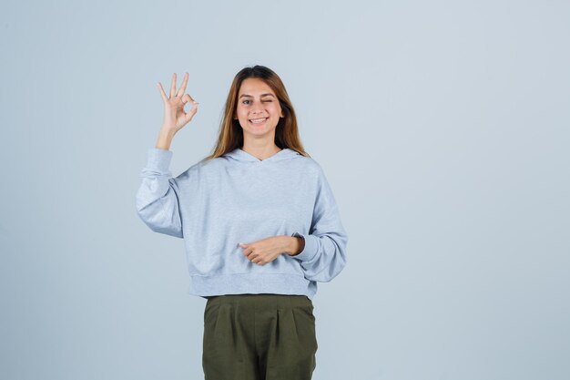 Blonde girl showing ok sign in olive green blue sweatshirt and pants and looking radiant , front view.