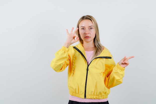확인 표시를 보여주는 금발 소녀, 분홍색 티셔츠와 노란색 재킷에 오른쪽을 가리키고 심각한 찾고.