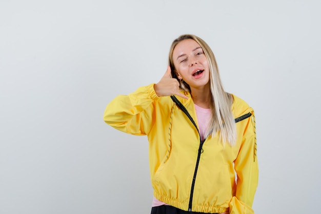Блондинка показывает телефонный жест в желтой куртке и выглядит уверенно.