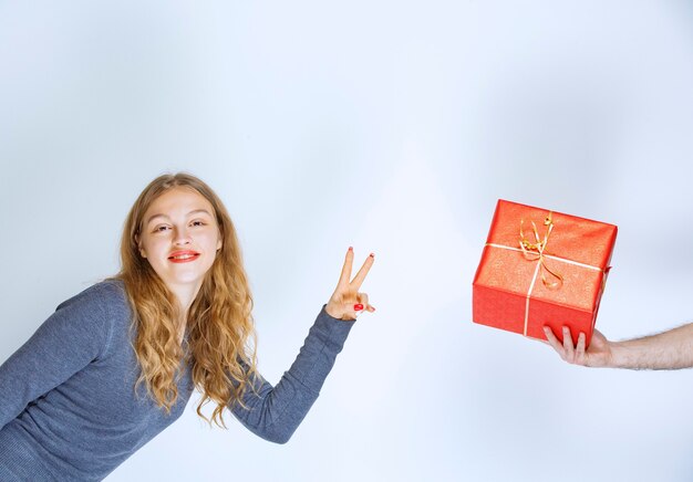 Blonde girl showing a red gift box and making peace sign.