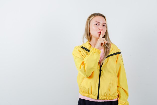 Blonde girl showing silence gesture in pink t-shirt and yellow jacket and looking alluring.