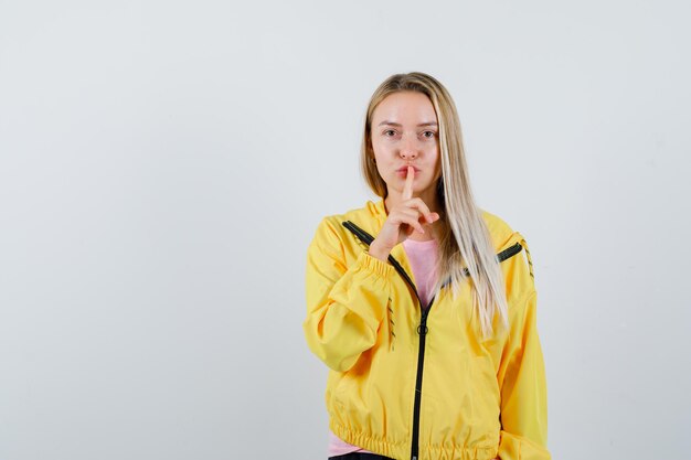 Блондинка показывает жест молчания в желтой куртке и выглядит разумно