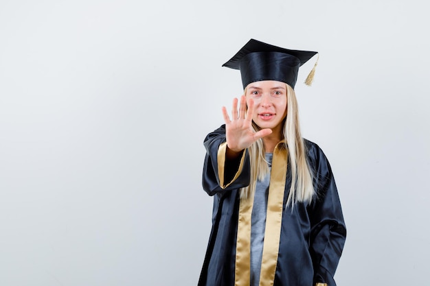 Blonde girl showing stop sign in graduation gown and cap and looking happy.