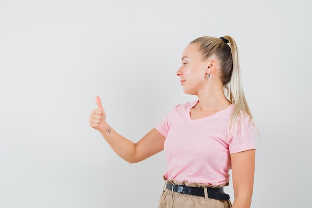 Ragazza bionda che mostra il pollice in su e sorridente in t-shirt, pantaloni vista frontale.