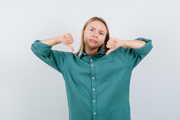 Free photo blonde girl showing thumbs down with both hands in green blouse and looking displeased.