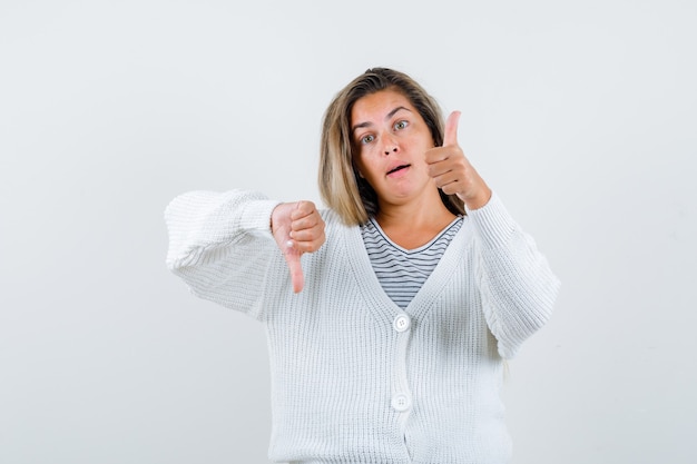 Blonde girl showing  thumbs up and down in striped t-shirt, white cardigan and jean pants and looking serious, front view.