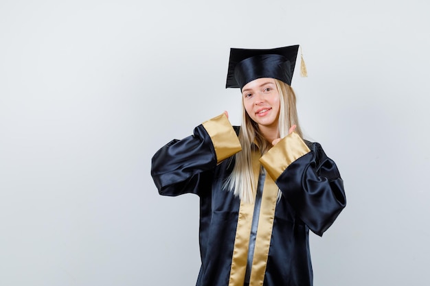 Blonde girl showing thumbs up with both hands in graduation gown and cap and looking cute.