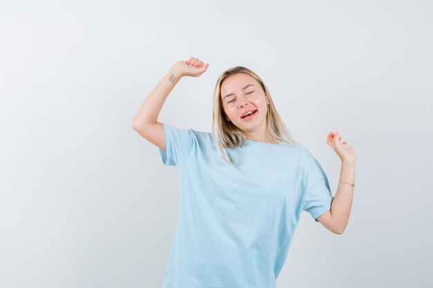 Блондинка показывает жест победителя в синей футболке и выглядит весело, вид спереди.
