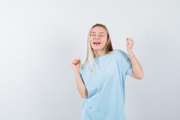 青いTシャツで勝者のジェスチャーを示し、幸運に見えるブロンドの女の子。正面図。