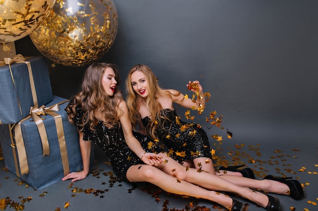 Blonde girl sitting on the floor with friend and throwing out golden confetti. Stylish ladies in black dresses lying beside presents and balloons and joking.