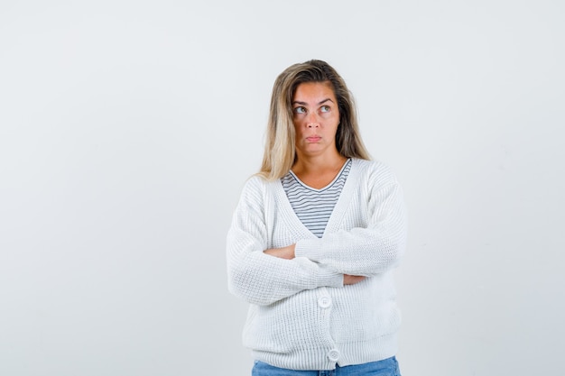 Blonde girl standing arms crossed in striped t-shirt, white cardigan and jean pants and looking pensive , front view.