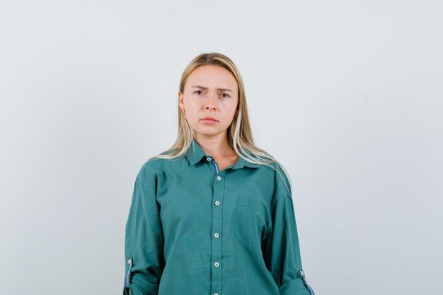 Blonde girl standing straight, grimacing and posing at camera in green blouse and looking displeased.