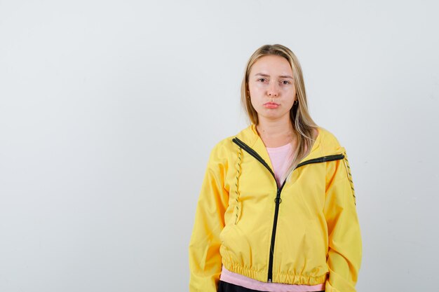Блондинка стоит прямо и позирует перед камерой в розовой футболке и желтой куртке и выглядит серьезно.