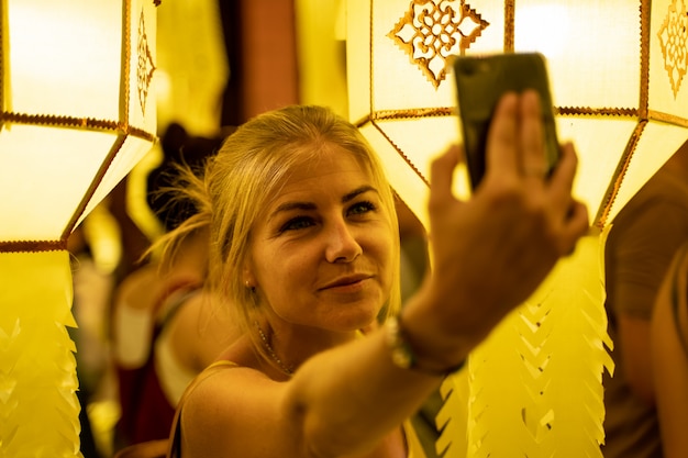 Blonde girl in a strapless dress surrounded by Chinese lanterns at night making a selfie