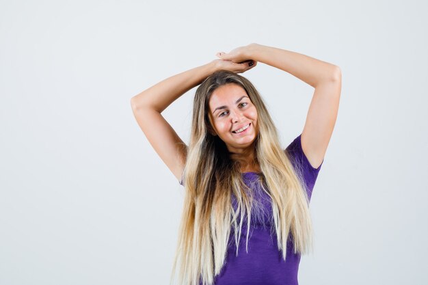 紫のTシャツで頭上に腕を伸ばしてリラックスして見えるブロンドの女の子、正面図。