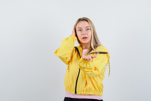 Blonde girl stretching hand to offer something in yellow jacket and looking cute.