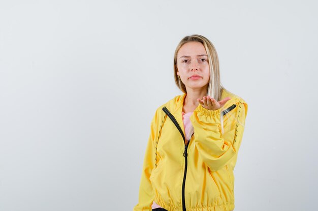 Blonde girl stretching hand in questioning gesture in yellow jacket and looking confused.