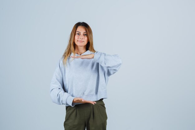 Blonde girl stretching hands as holding something in olive green blue sweatshirt and pants and looking cheery , front view.