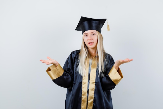 Free photo blonde girl stretching hands in questioning manner in graduation gown and cap and looking perplexed