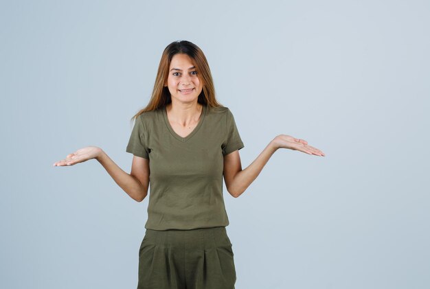 Blonde girl stretching hands in questioning manner in olive green t-shirt and pants and looking baffled. front view.