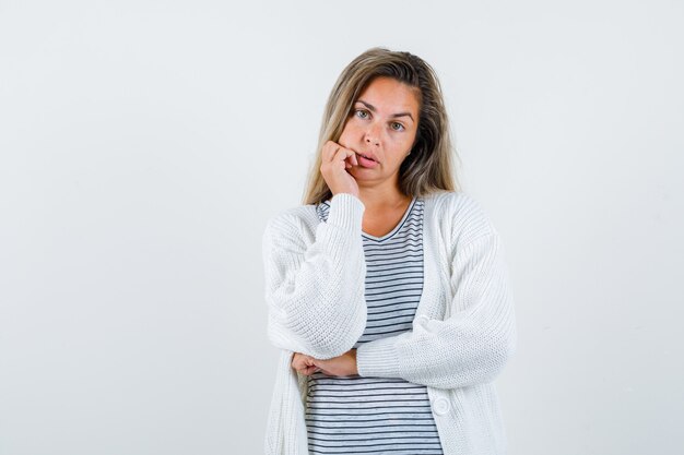 Blonde girl in striped t-shirt, white cardigan and jean pants leaning chin on palm and looking pensive , front view.