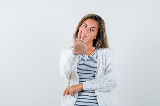 ストライプのTシャツ、白いカーディガン、ジーンズのズボンのブロンドの女の子は一時停止の標識を示し、真剣に見える、正面図。